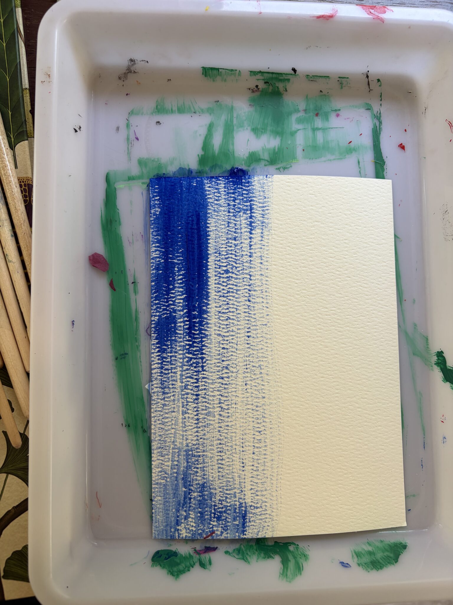 Close up of a painting in progress showing a light vertical wash of blue paint drying on raw textured white paper.