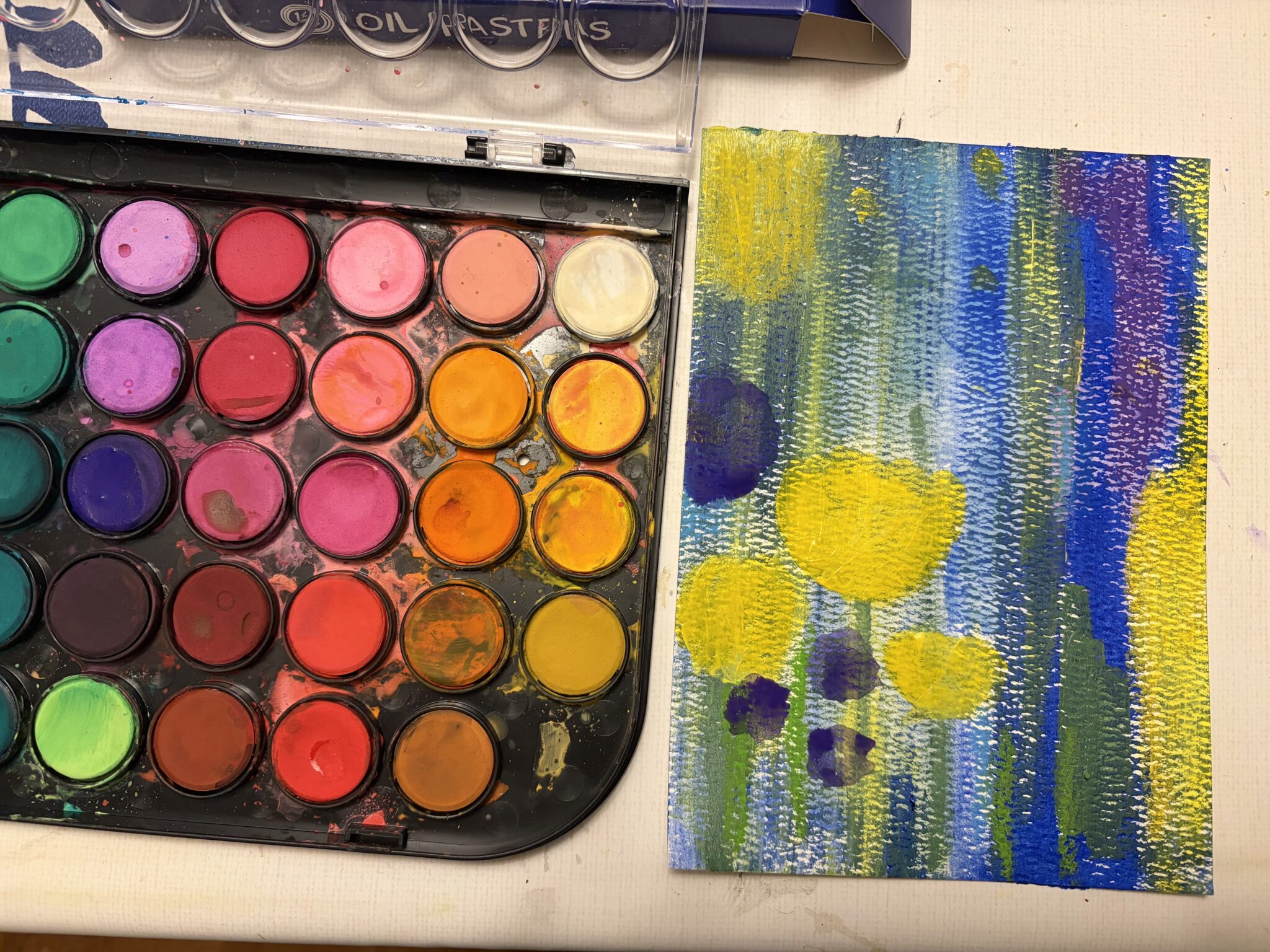 A colorful watercolor paint palette next to blue and yellow acrylic wash paintinh